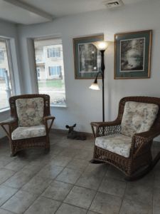 A cozy living room corner featuring two rattan chairs with floral cushions, a floor lamp, and framed artwork on the walls.