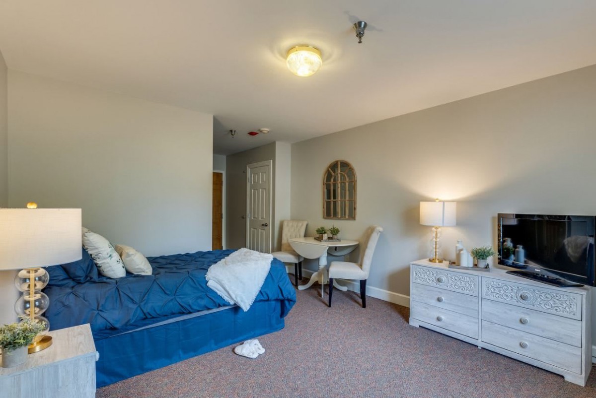 A cozy bedroom with a blue bed, table and chairs, a lamp on a dresser, and soft lighting.