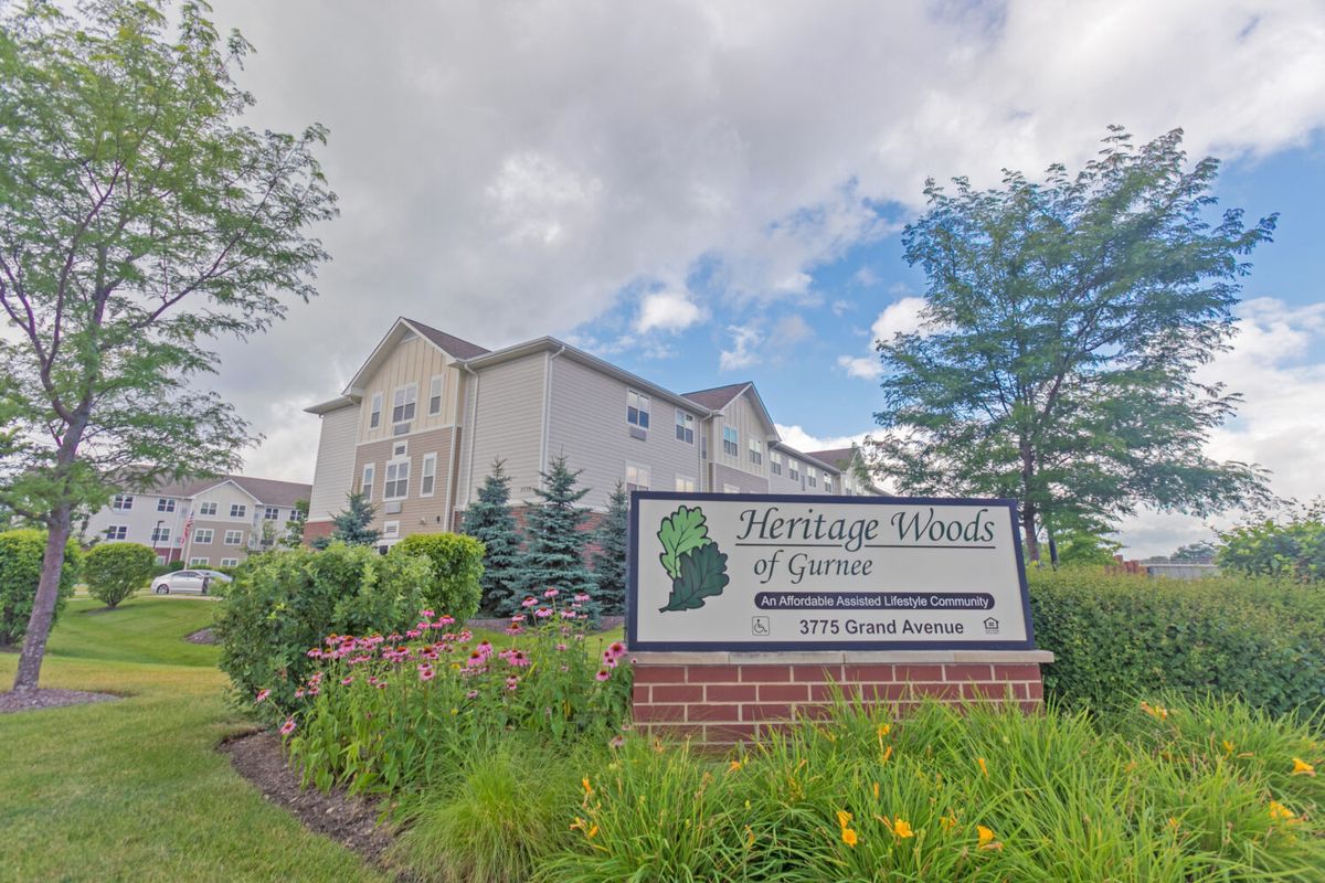 A multi-story building with a sign for Heritage Woods of Batavia, set amidst a landscaped area and under a cloudy sky.