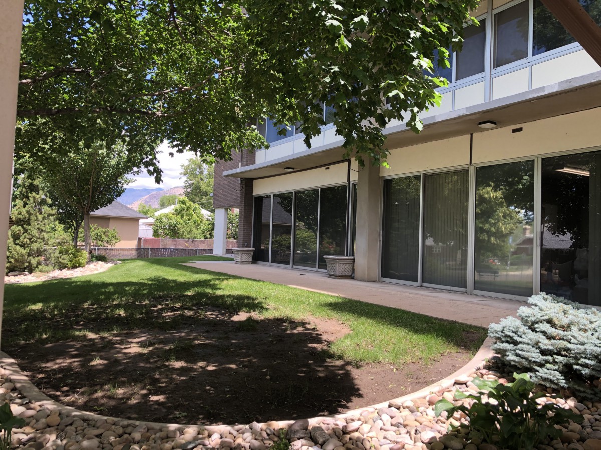 A modern building with a patio, surrounded by a well-maintained lawn and trees on a sunny day.