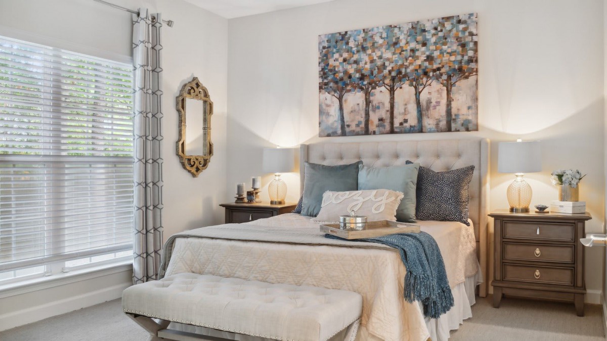 A cozy and elegantly designed bedroom featuring a bed with multiple pillows, a soft blanket, an artistic wall piece above the headboard, a decorative mirror, and a nightstand with a lamp.