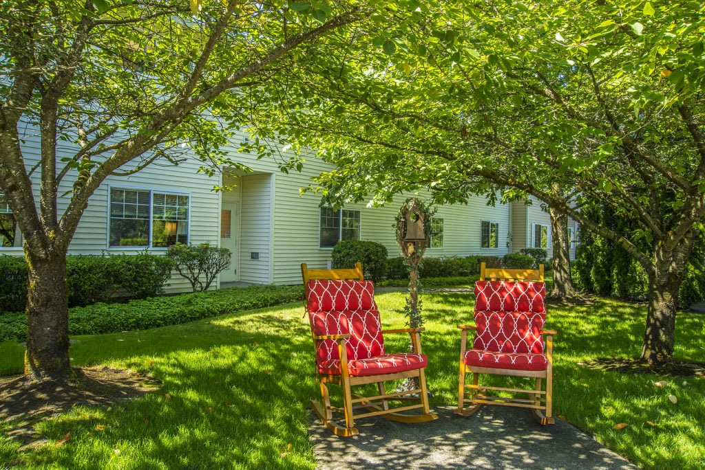 A peaceful garden setting with two red rocking chairs under a tree, in front of a white house.