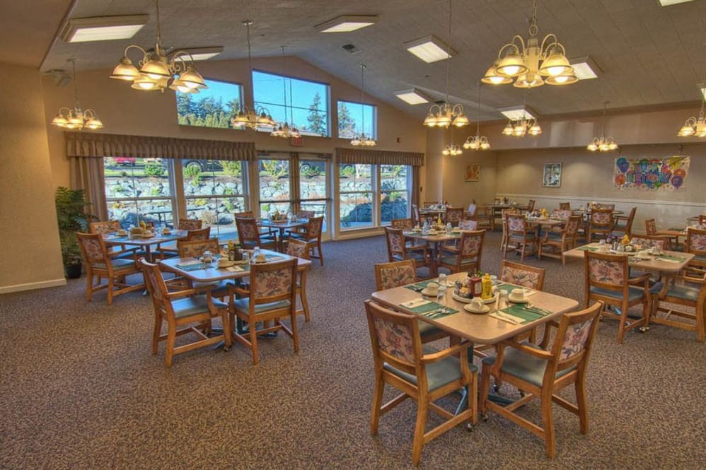 A cozy restaurant dining area with neatly arranged tables and chairs, illuminated by chandeliers and natural light from large windows.