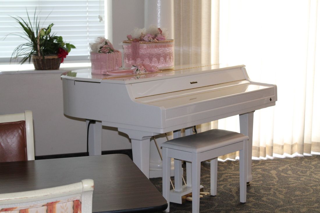 A white piano with a matching bench, adorned with pink floral decorations, situated in a sunlit room.