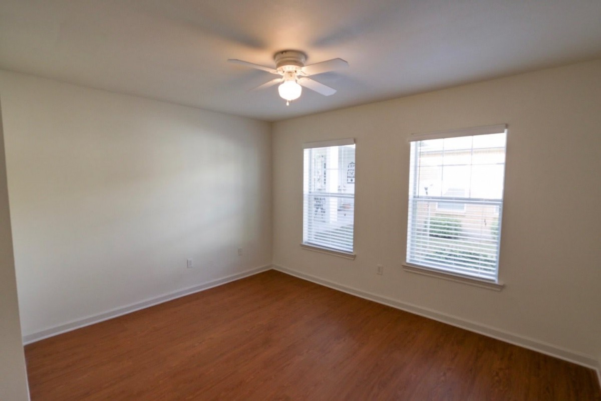 An empty room with wood flooring, two windows letting in natural light, and a ceiling fan.