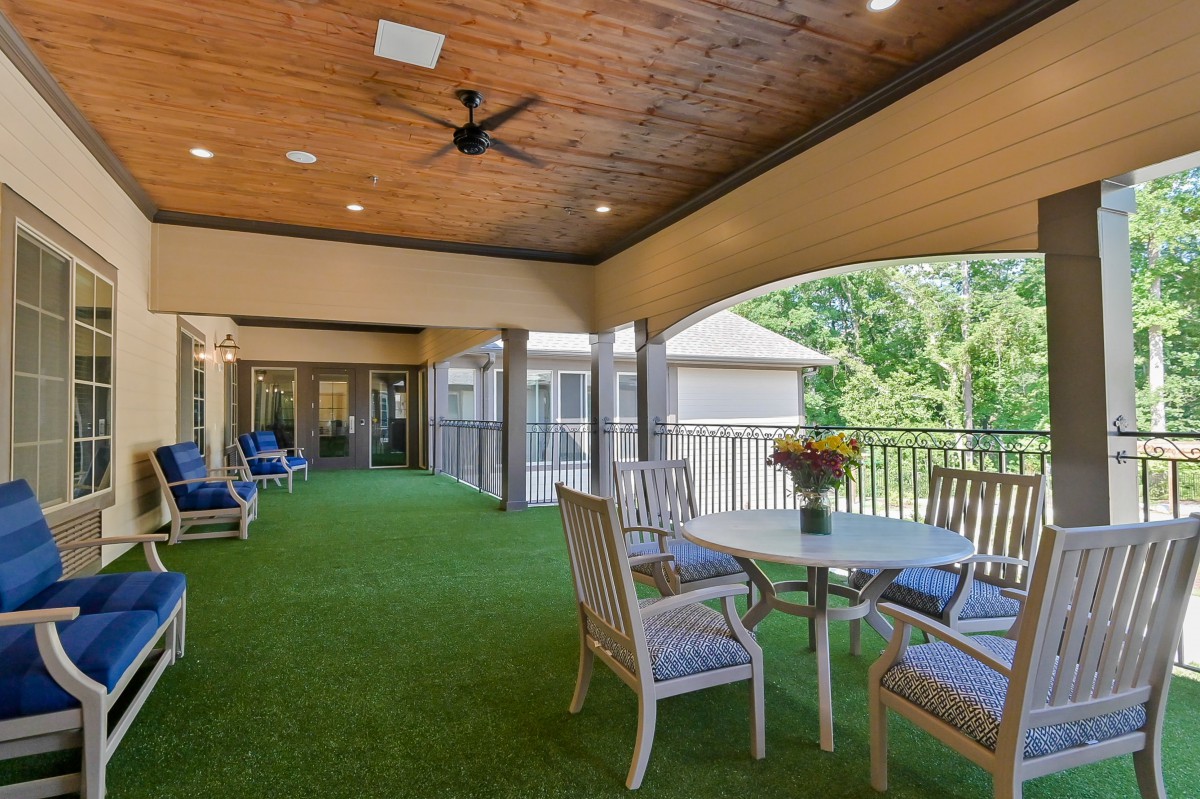 A cozy covered patio with artificial grass, featuring a wooden ceiling, ceiling fan, outdoor seating, and a table with flowers.