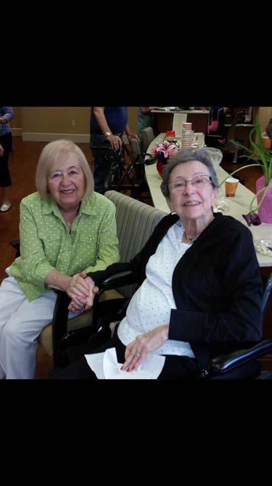 Two elderly women smiling and holding hands indoors.