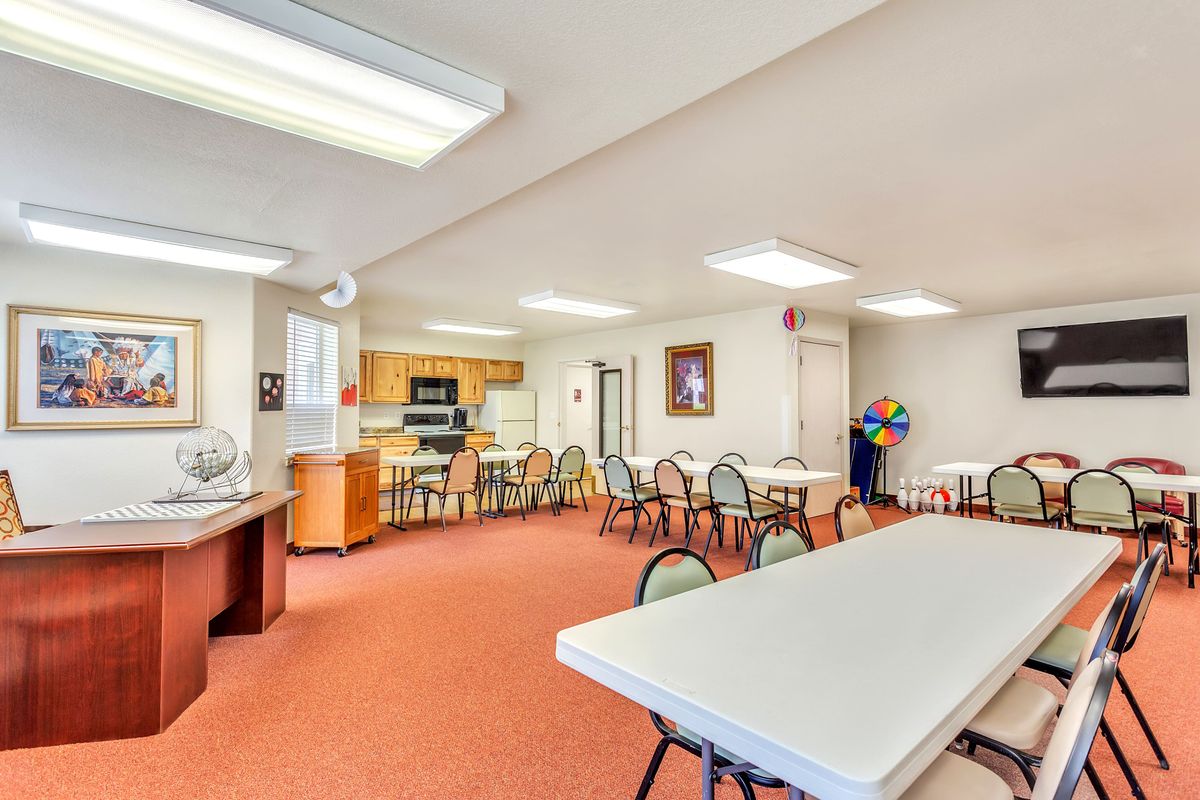 A spacious community room with tables, chairs, and a kitchen area under bright indoor lighting.