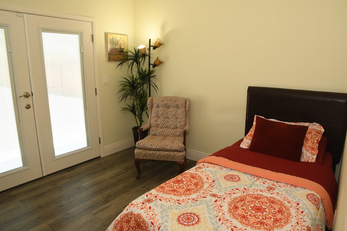 A cozy bedroom with a decorative bedspread, an armchair, a floor lamp, and a potted plant next to a glass door.