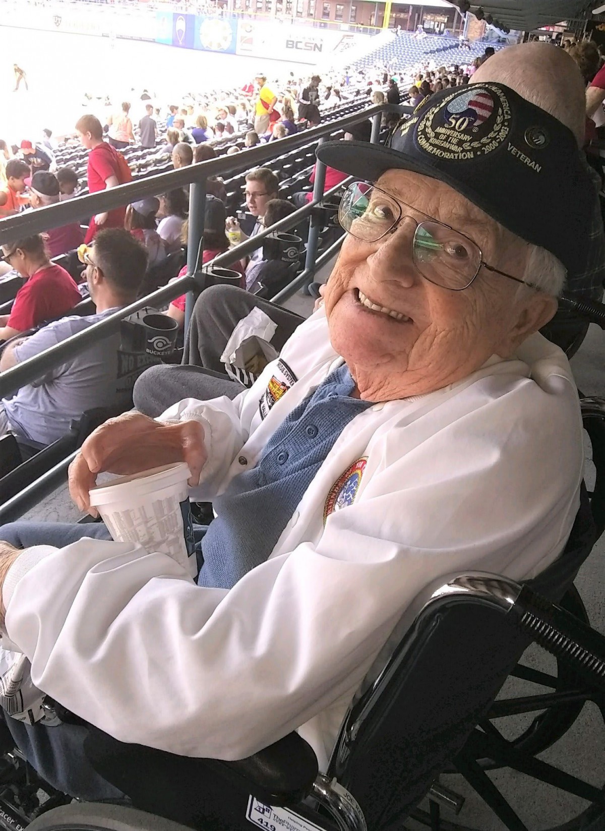 An elderly man wearing a baseball cap and glasses, smiling while seated in a stadium.