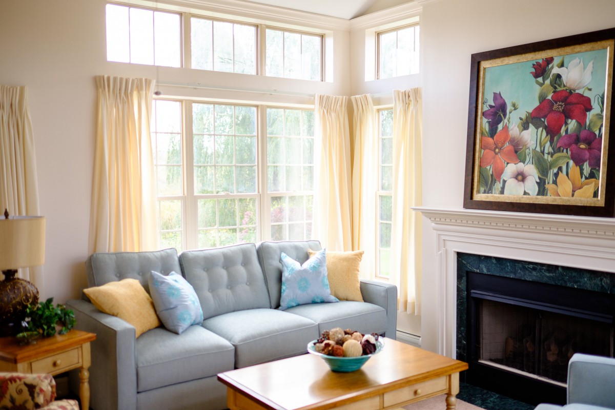 A cozy living room with a light blue sofa, accented by yellow and blue cushions. Large windows with sheer curtains allow natural light to fill the space. A wooden coffee table sits in front of the sofa, topped with a decorative bowl. The room features a f