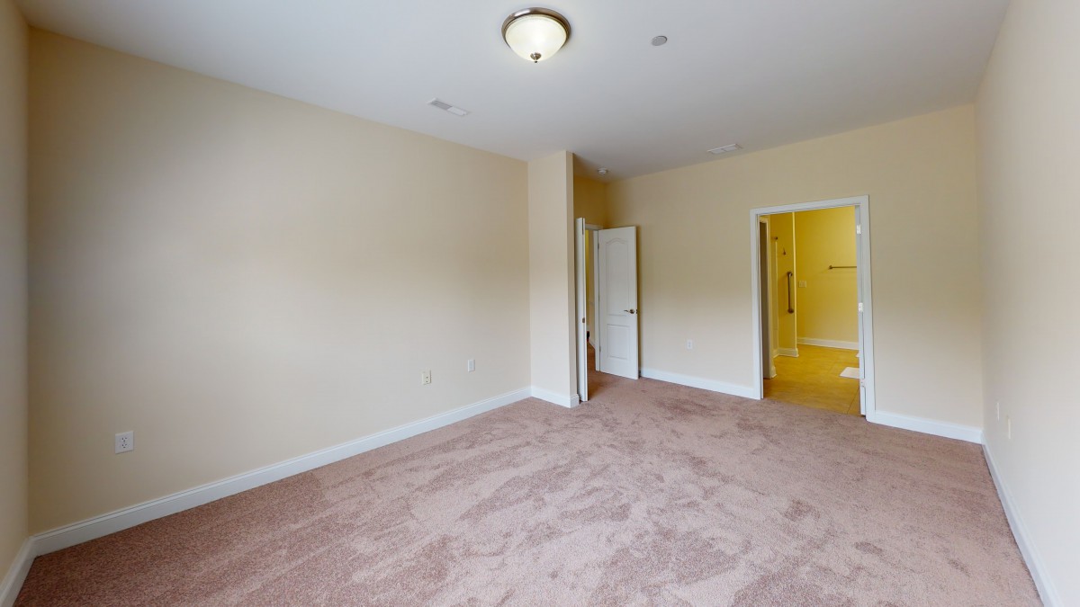 An empty room with beige walls, a carpeted floor, and a ceiling light. An open door leads to another space.