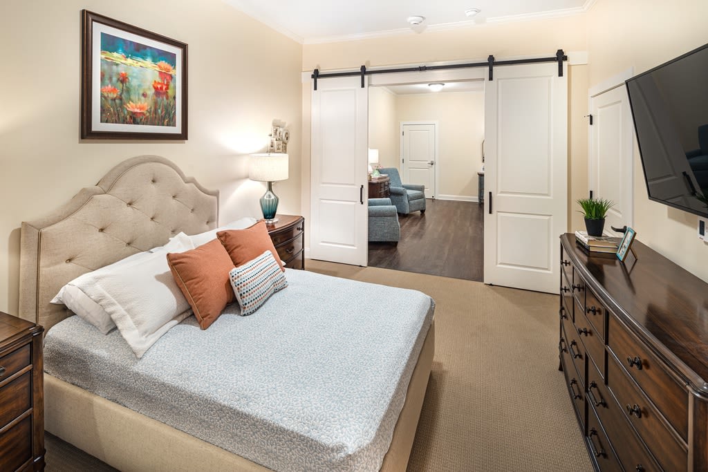 A cozy bedroom with a neatly made bed, featuring colorful pillows and artwork, separated from a sitting area by sliding doors.