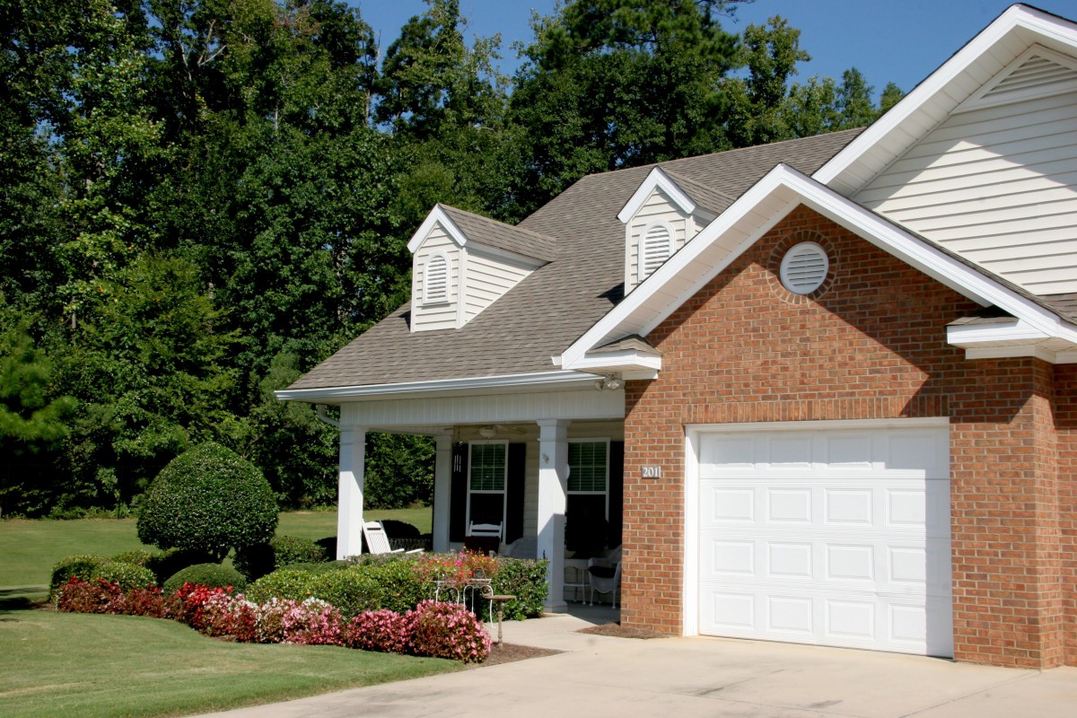 A suburban brick house with a well-maintained lawn and garden, featuring a garage and covered porch.