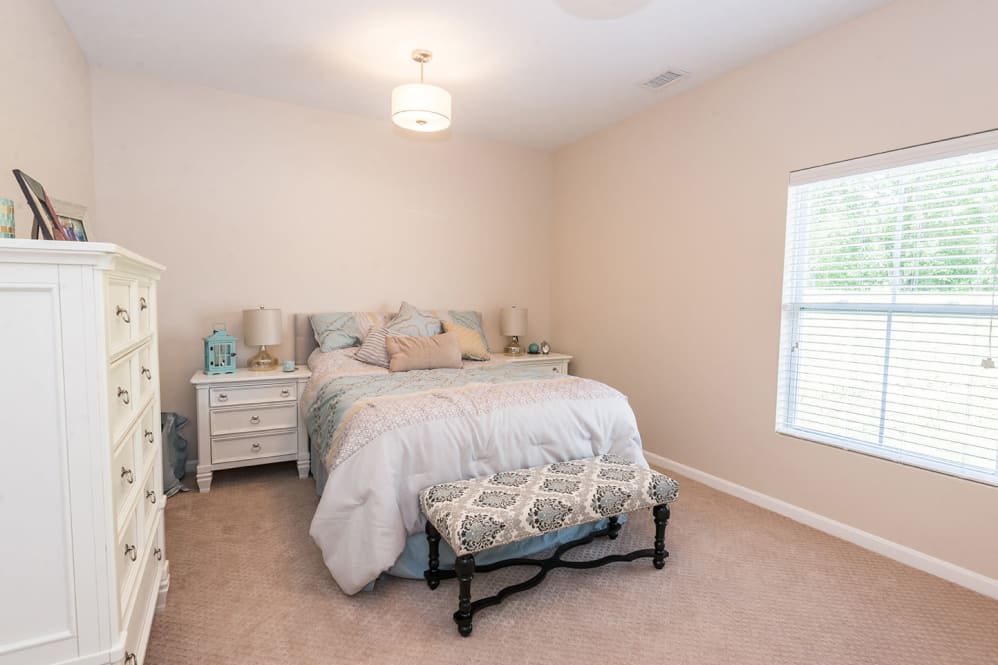 A cozy bedroom with a neatly made bed, nightstand, and a window allowing natural light.