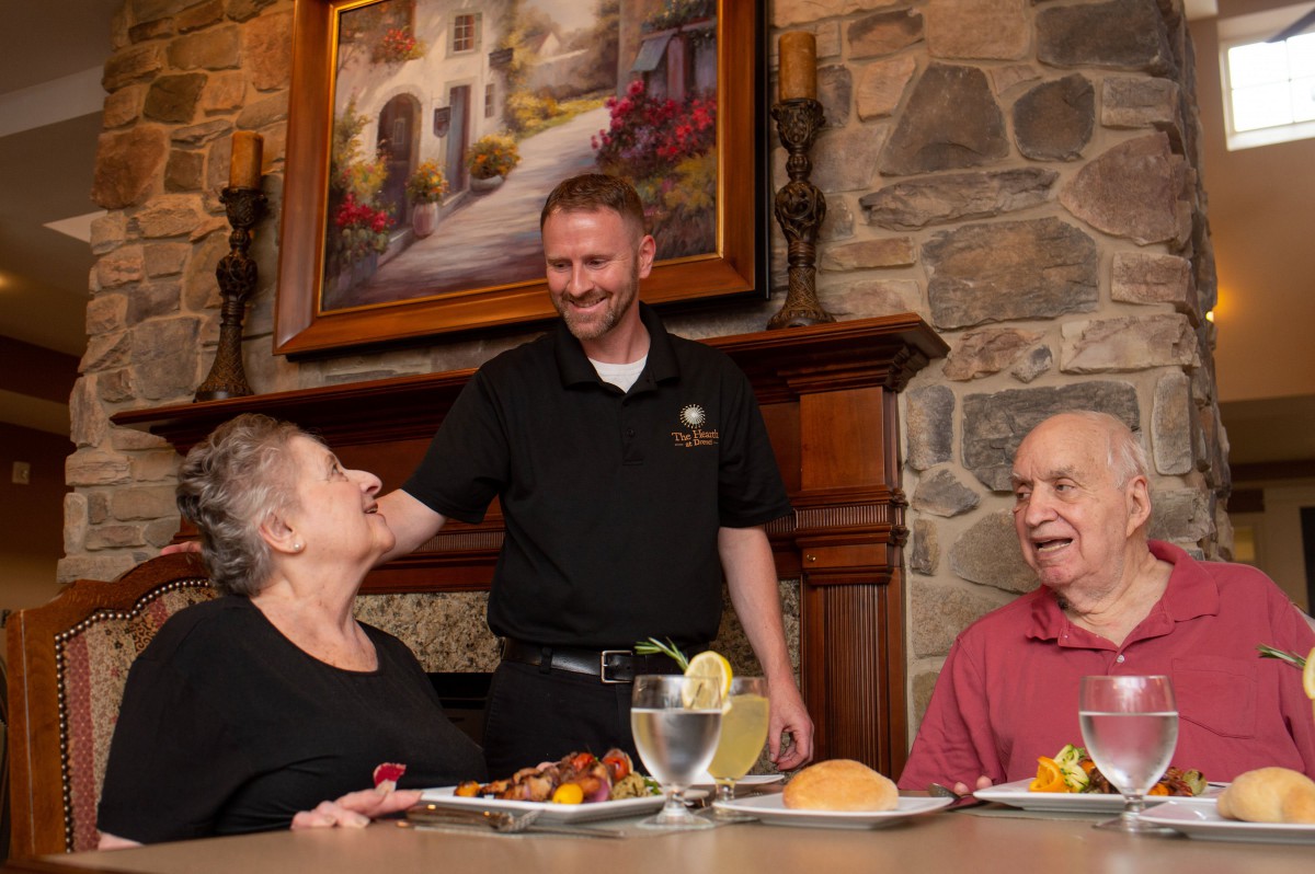 An elderly couple enjoys a meal at a restaurant, while a friendly waiter engages in conversation.