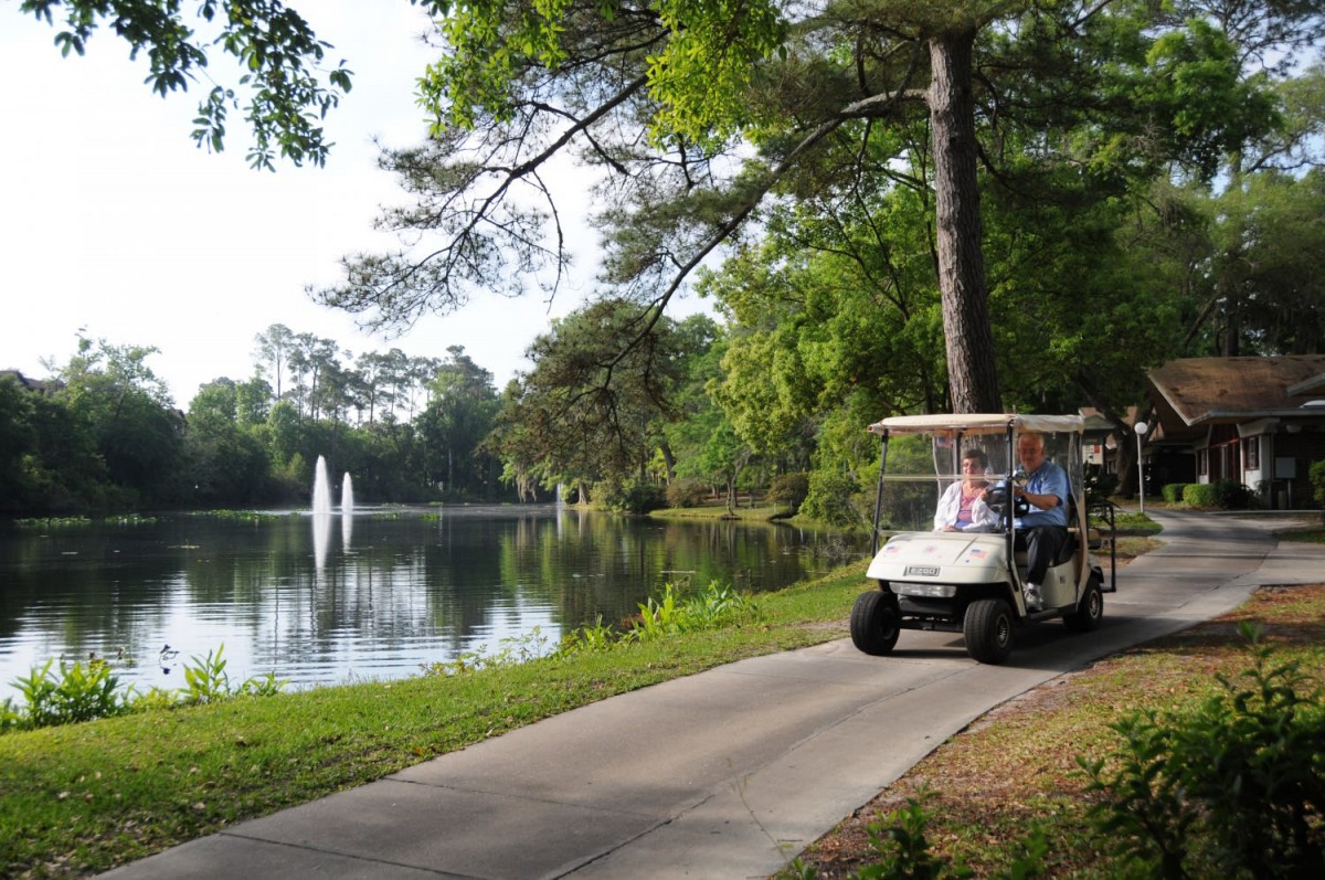 A golf cart driving along a scenic pathway by the lake, surrounded by trees and a fountain.