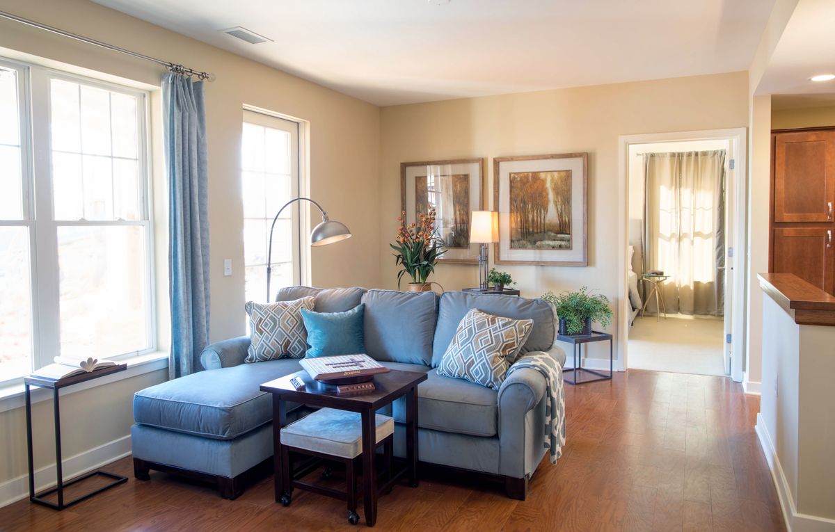 A cozy living room with a blue sectional sofa, wooden flooring, and natural light streaming through large windows.