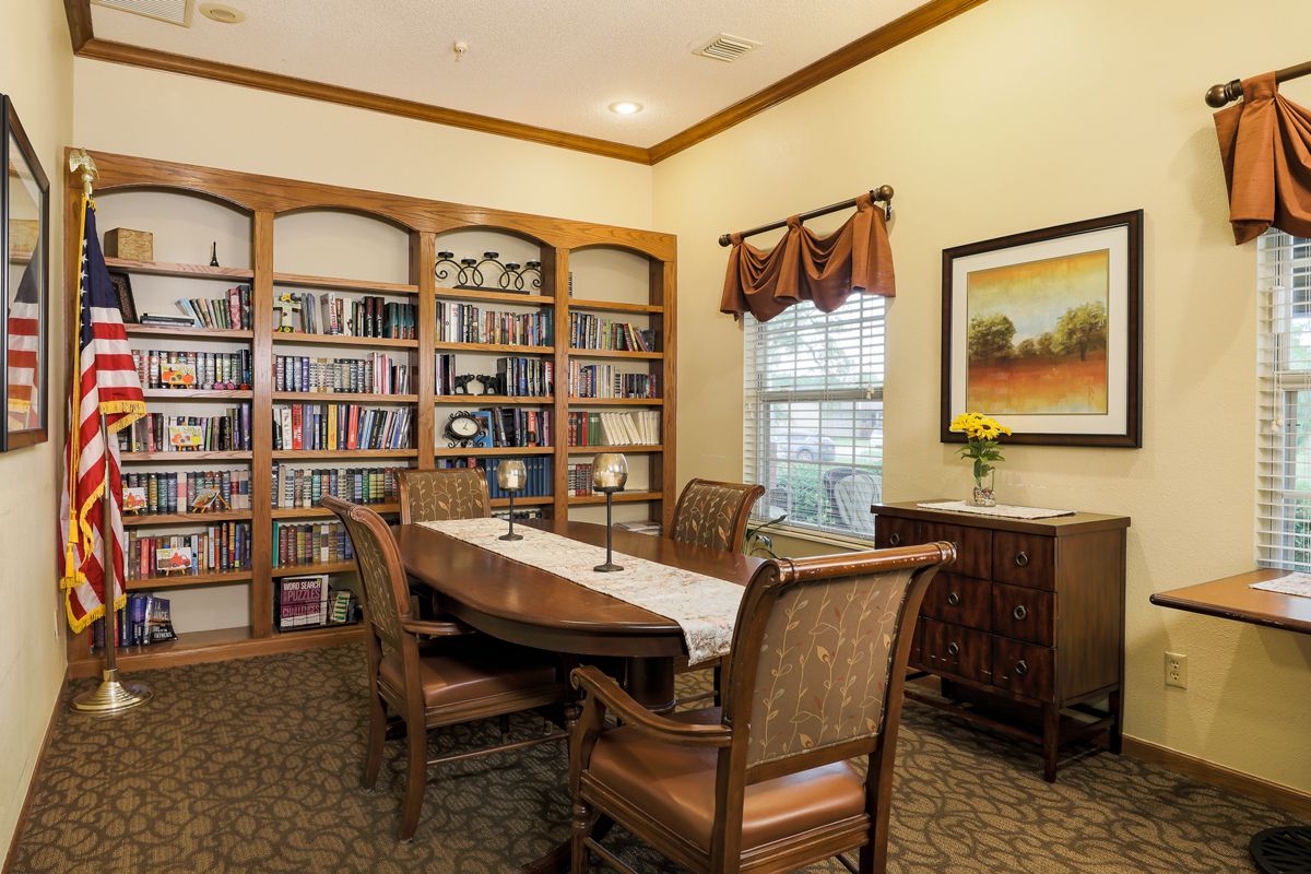 A cozy library room featuring a large bookshelf, a table with chairs, and a window with curtains.