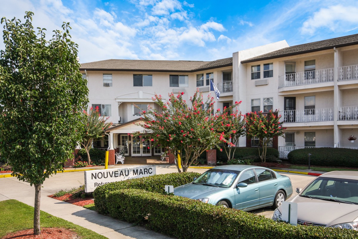 A multi-story apartment building with a car parked in front and trees lining the entrance.