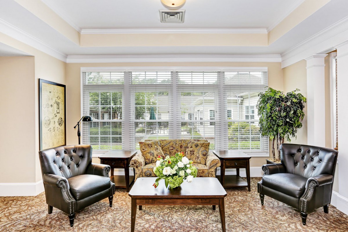 A cozy living room with large windows, featuring comfortable armchairs, a patterned sofa, and a coffee table adorned with a floral arrangement.