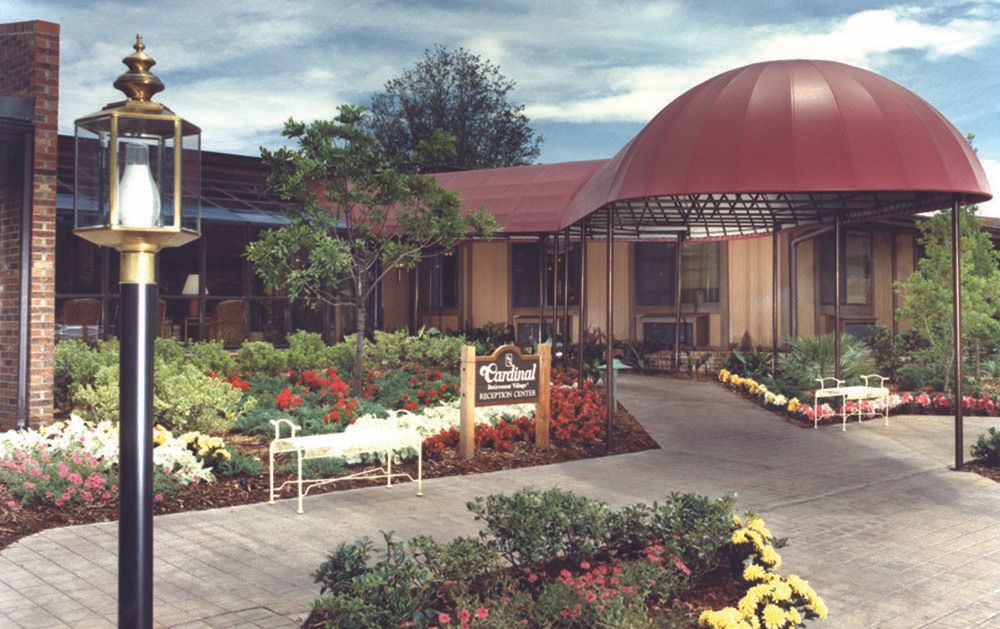 A scenic garden with a variety of plants and flowers in front of a building with a red domed roof, featuring a pathway and bench.