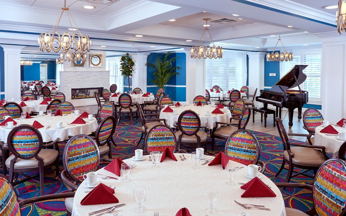 An elegantly decorated dining room with round tables set with red napkins and colorful chairs, featuring a piano in the corner.