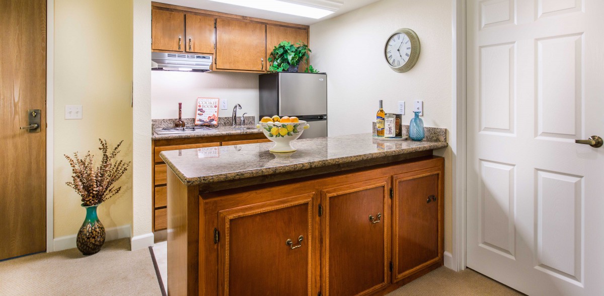 A cozy kitchen with wooden cabinets and a granite countertop, featuring an island centerpiece adorned with fresh fruits and wine bottles.