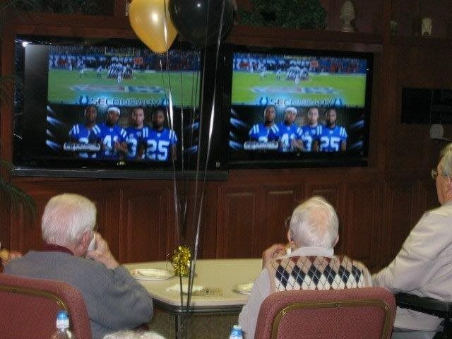 A group of seniors watching a football game on two televisions, with balloons celebrating the occasion.