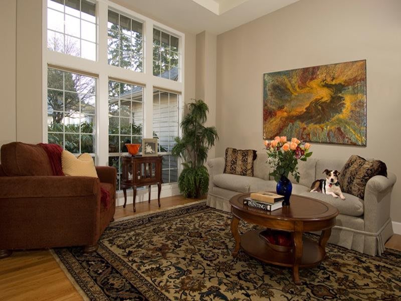 A cozy living room with large windows, a plush sofa, a wooden coffee table adorned with flowers, and vibrant artwork on the wall.