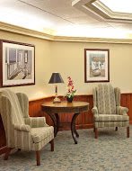 A cozy lobby area with two upholstered chairs, a round table with a lamp and a pink flower, all set against wood-paneled walls with framed pictures.