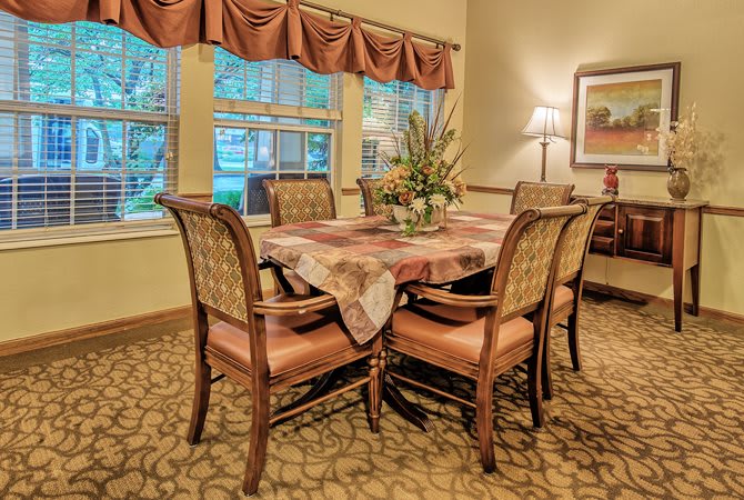 A cozy dining room with a set table and upholstered chairs, framed by large windows and warm lighting.