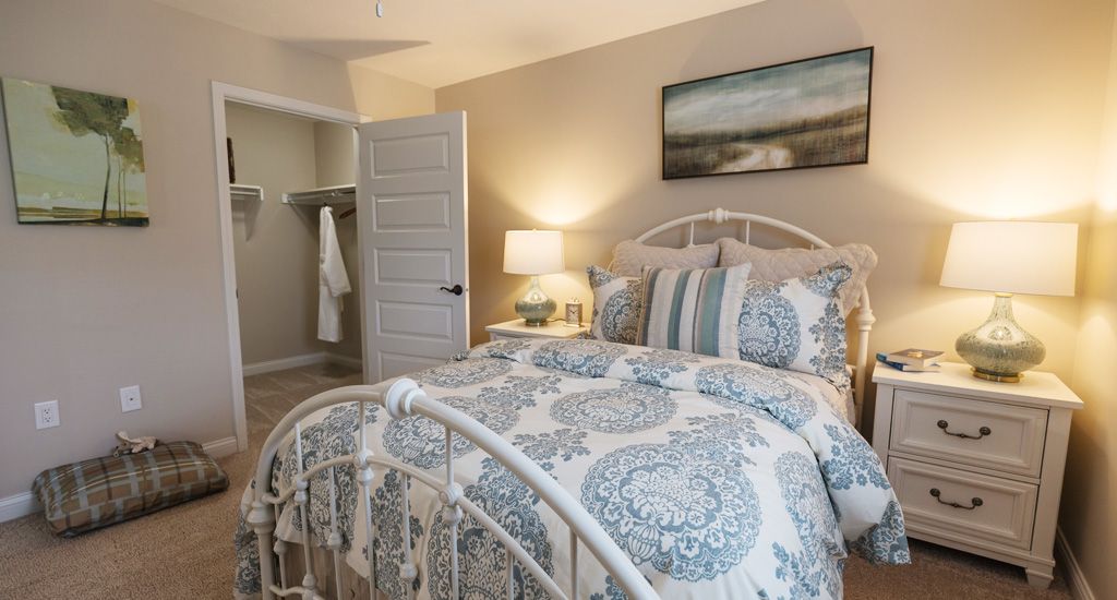 A cozy bedroom interior featuring a well-decorated bed with patterned bedding, a nightstand with a lamp, and artwork on the wall.