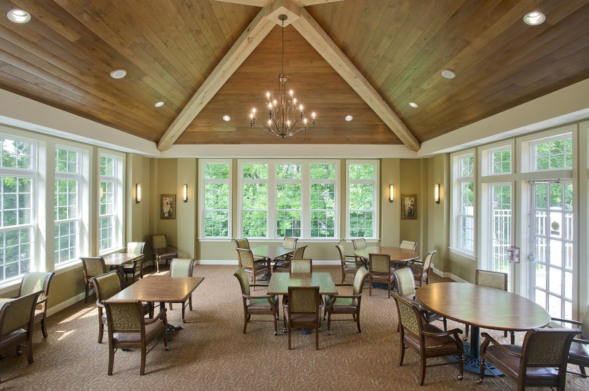 A spacious dining room with several tables and chairs, illuminated by a chandelier and natural light streaming through large windows.