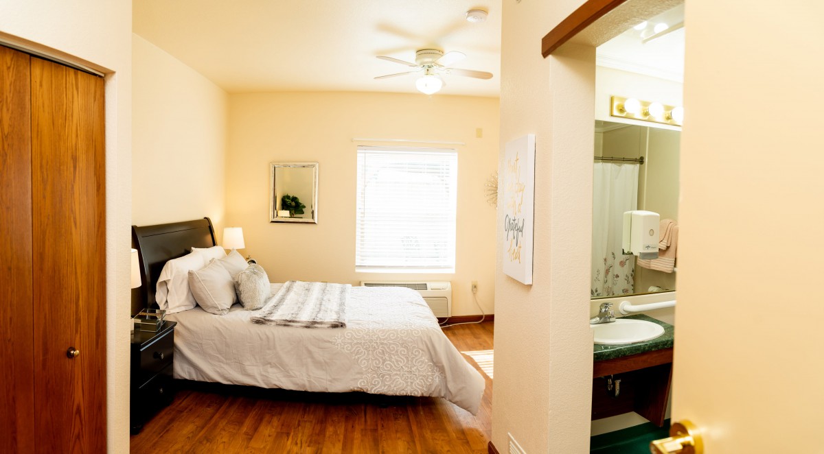 A cozy bedroom with a neatly made bed, hardwood floors, and an adjoining bathroom.