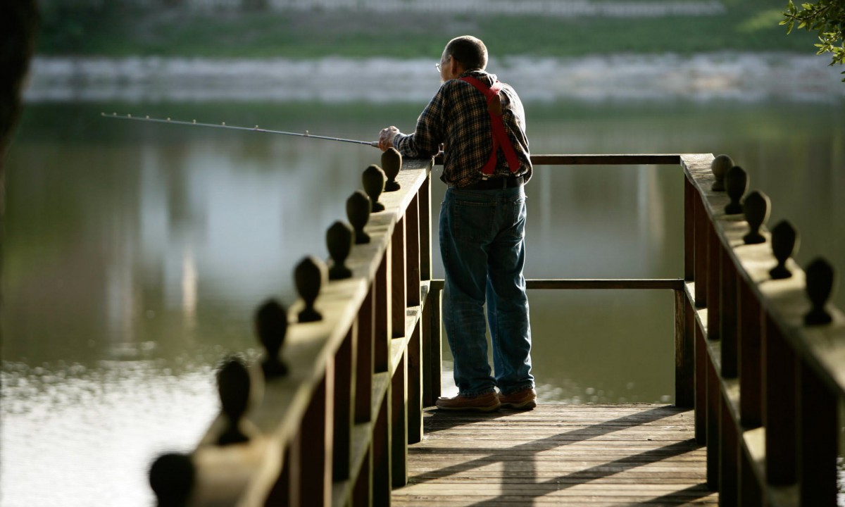 A man is fishing while standing on a dock by the lake.