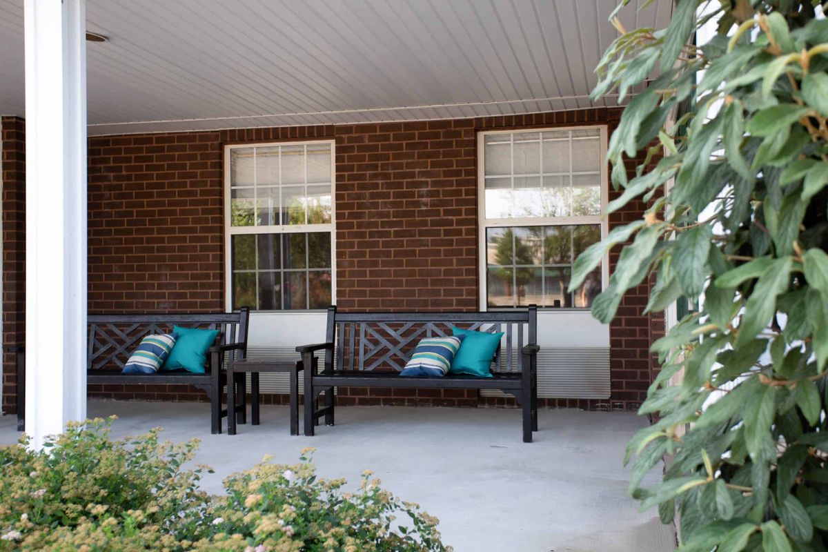 A cozy porch featuring two benches adorned with colorful cushions alongside a brick wall and windows.