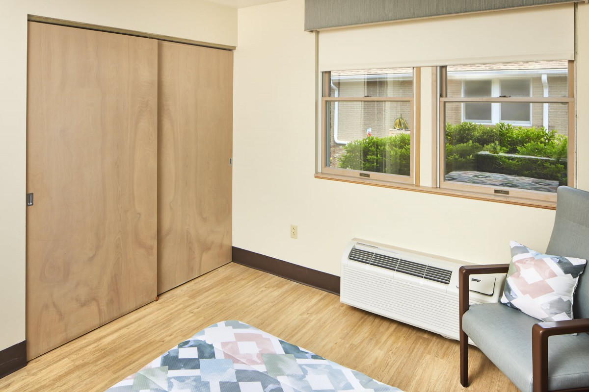 A minimalist bedroom with a wooden floor, featuring a closet, window with natural light, and an air conditioning unit.