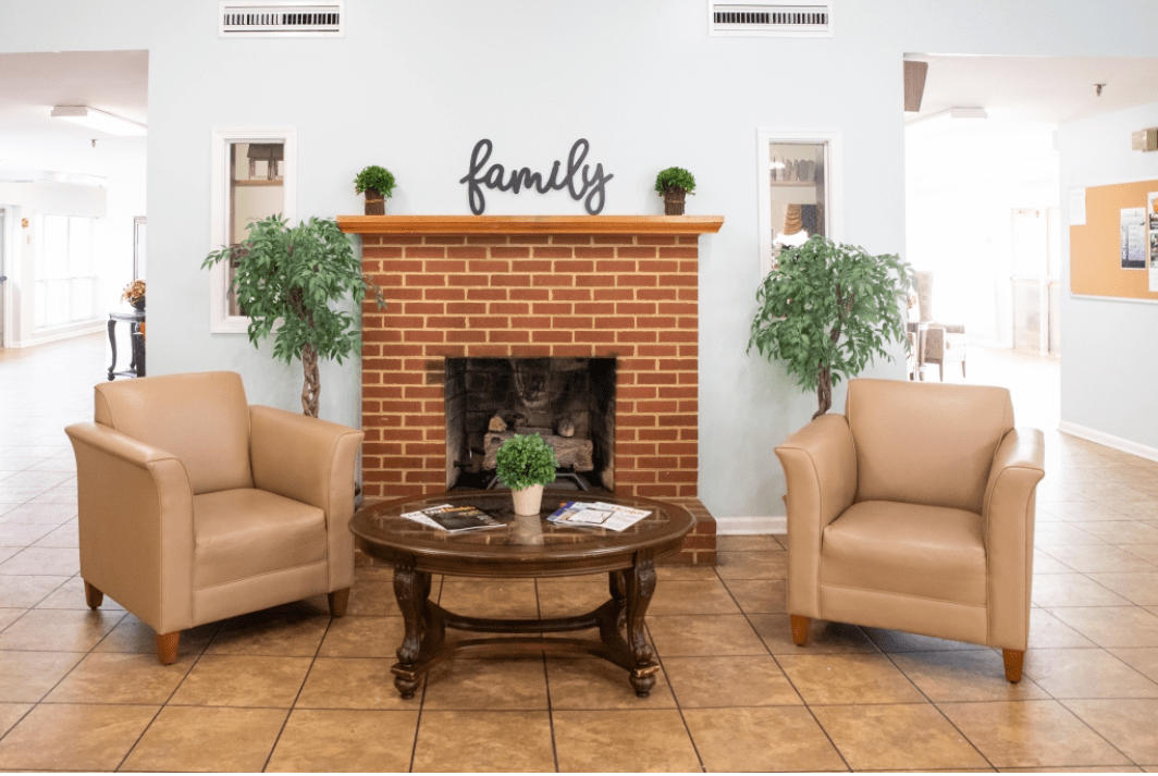 A cozy living room with a brick fireplace, two beige chairs, green indoor plants, and a wooden coffee table.