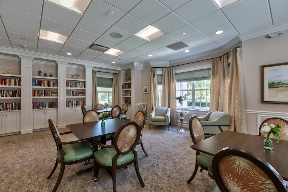 A spacious, well-lit room featuring multiple tables and chairs, with bookshelves lining the walls and large windows adorned with curtains.