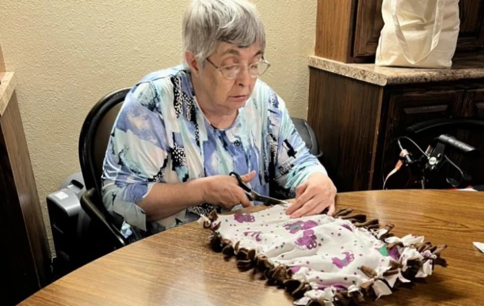 An elderly woman is focused on crafting, cutting fabric at a table.