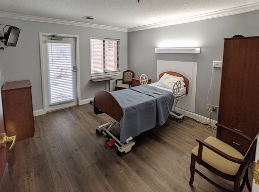 A simple and well-lit hospital patient room featuring a bed, a chair, and windows for natural light.