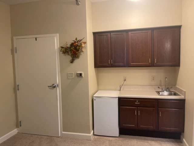 A simple kitchen area with dark cabinets, a small refrigerator, and a sink next to a door.