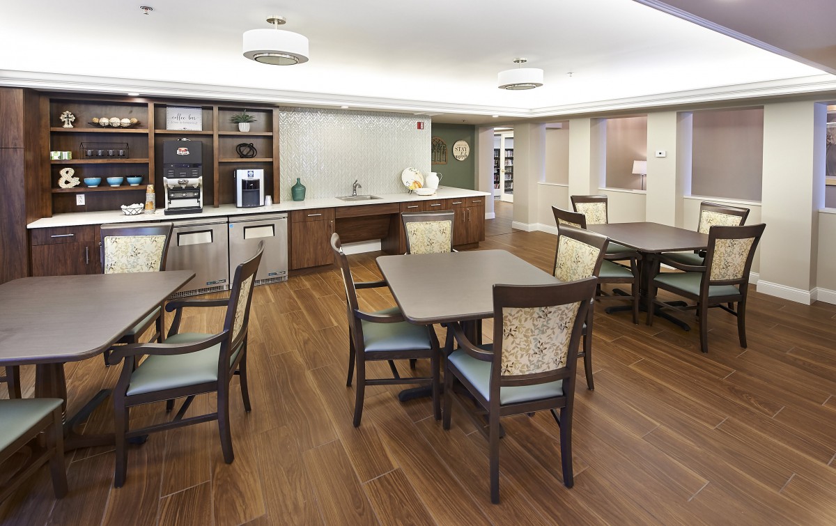 A modern dining room with wooden flooring, featuring multiple tables and chairs, and a kitchen area in the background.