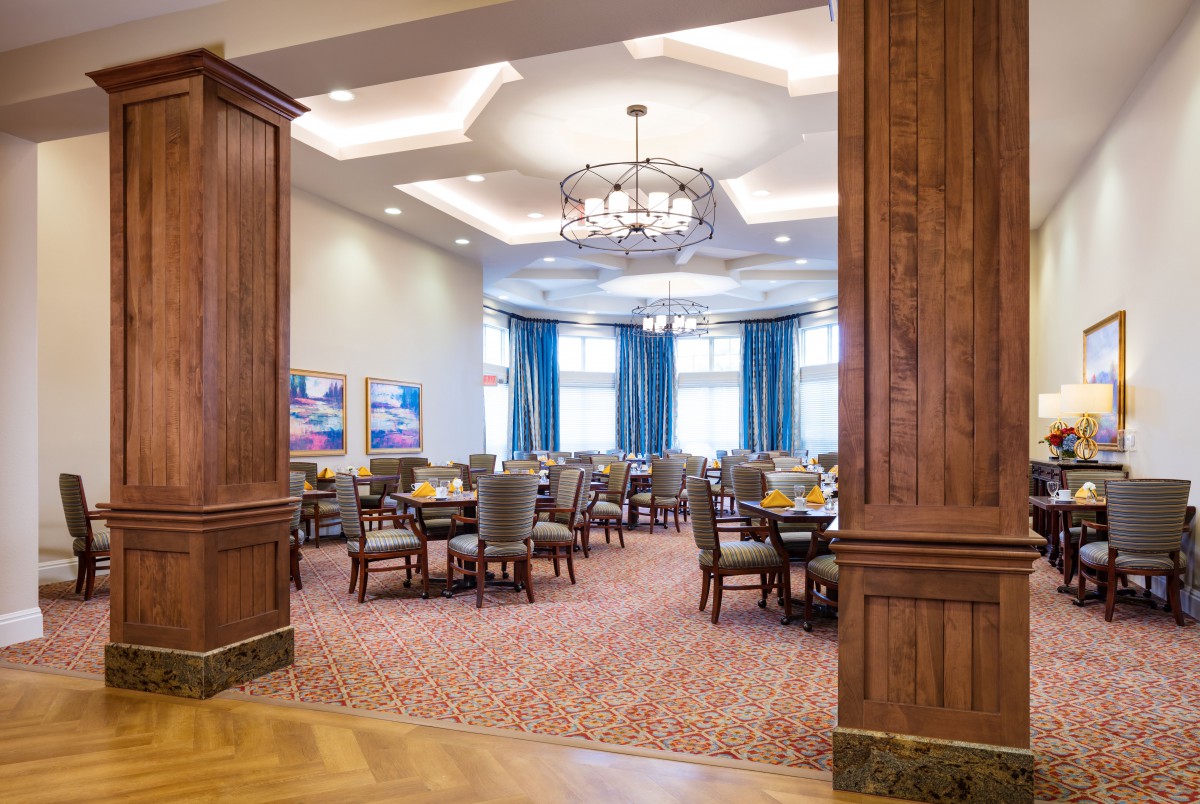 Elegant dining room with neatly arranged tables and chairs, featuring chandeliers and large windows with blue curtains.