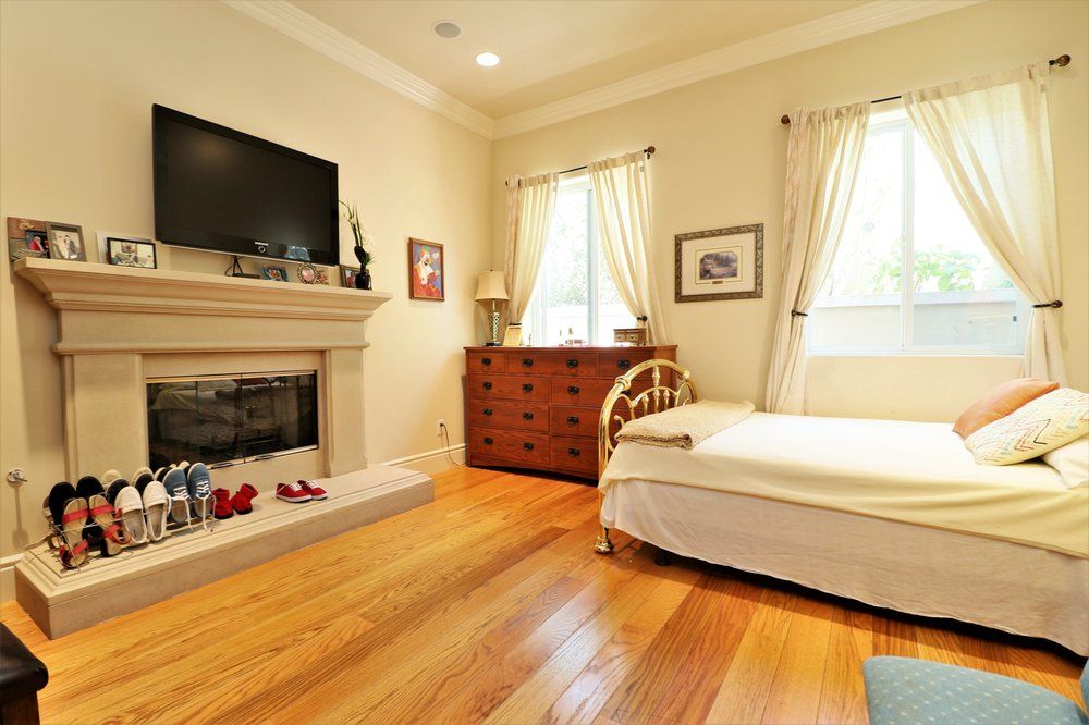 Cozy bedroom with a fireplace and television, featuring wooden flooring and natural light from two windows.