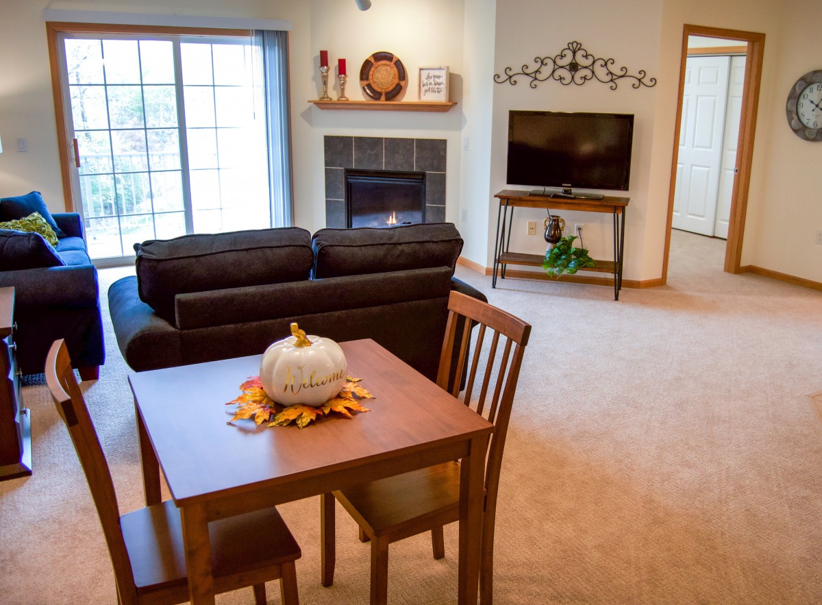 A cozy living room with a dining area features a brown sofa, a wooden table with a decorative pumpkin centerpiece, and a TV set next to a fireplace.