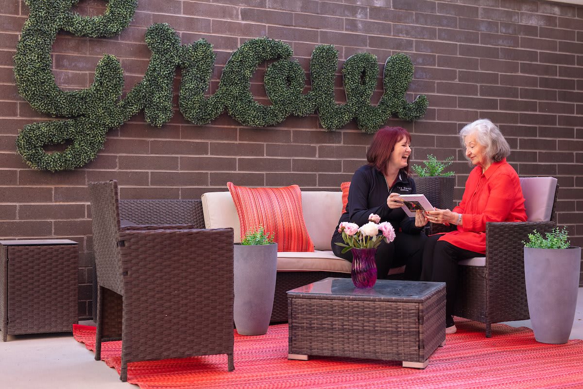 Two women enjoy a conversation in an outdoor seating area with rattan furniture and greenery.