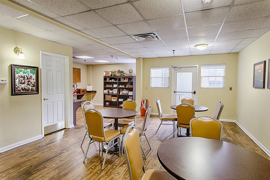 A community room with round tables and chairs set on a wooden floor, featuring windows and a shelving unit in the background.