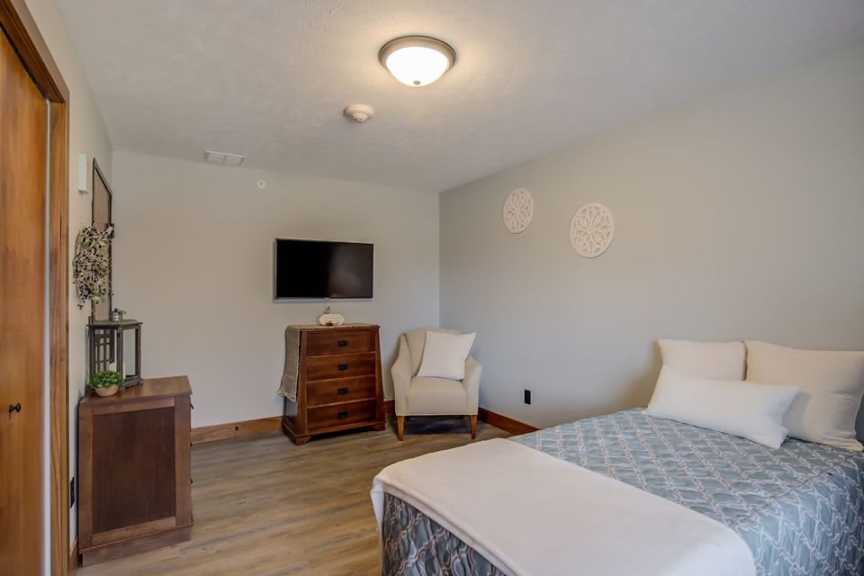 A minimalist bedroom with a bed, dresser, chair, and wall-mounted television.